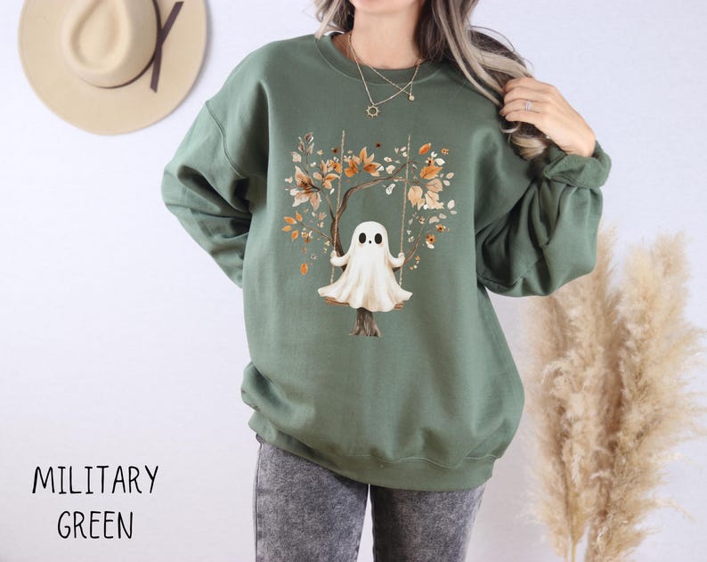 May include: Military green sweatshirt featuring a whimsical illustration of a ghost on a swing, set against a tree with autumn-colored leaves. The text "MILITARY GREEN" is printed at the bottom. The sweatshirt has long sleeves and a crew neck.