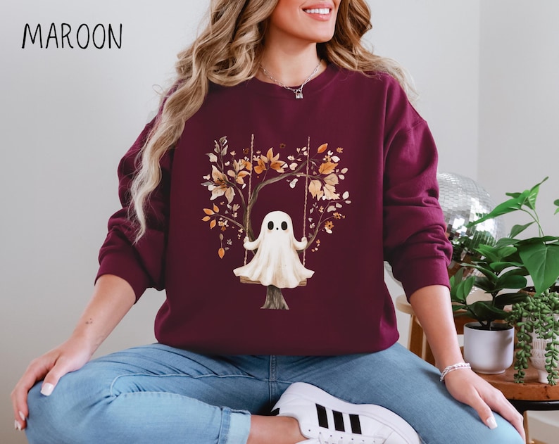 May include: Maroon sweatshirt featuring a whimsical illustration of a ghost on a swing, surrounded by autumn leaves. The text "MAROON" is printed at the top left. The design is centered on the front of the long-sleeved crew neck.