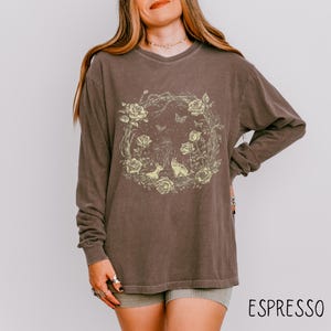 May include: Espresso-colored long-sleeve shirt featuring a floral wreath design with roses, butterflies, and woodland creatures. The shirt has a crew neck and a relaxed fit. The word "ESPRESSO" is printed in white at the bottom.