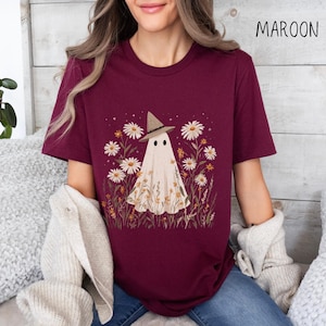 K&ouml;nnte beinhalten: Burgunderfarbenes T-Shirt mit einem Geist, der einen Hexenhut tr&auml;gt, umgeben von G&auml;nsebl&uuml;mchen und Wildblumen. Das Wort "MAROON" ist oben rechts in Schwarz aufgedruckt. Ein bequemes, l&auml;ssiges Oberteil f&uuml;r Liebhaber einzigartiger, von der Natur inspirierter Designs.