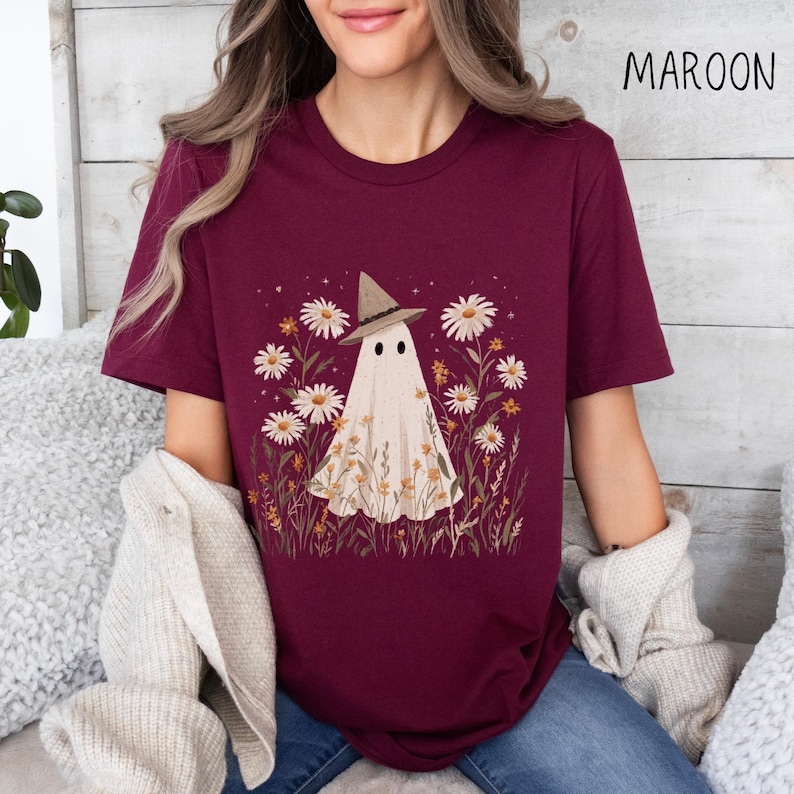 K&ouml;nnte beinhalten: Burgunderfarbenes T-Shirt mit einem Geist, der einen Hexenhut tr&auml;gt, umgeben von G&auml;nsebl&uuml;mchen und Wildblumen. Das Wort "MAROON" ist oben rechts in Schwarz aufgedruckt. Ein bequemes, l&auml;ssiges Oberteil f&uuml;r Liebhaber einzigartiger, von der Natur inspirierter Designs.