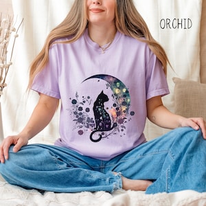May include: Orchid-colored t-shirt featuring a black cat silhouette seated on a crescent moon, surrounded by floral and botanical elements. The design includes butterflies and a colorful, celestial background. The word "ORCHID" is printed in black on the upper right.