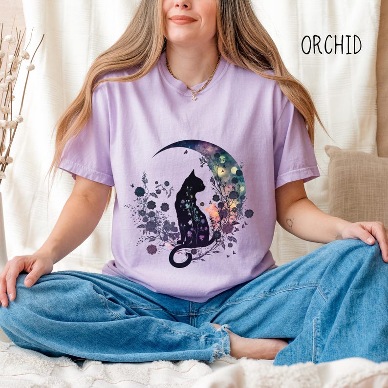 May include: Orchid-colored t-shirt featuring a black cat silhouette seated on a crescent moon, surrounded by floral and botanical elements. The design includes butterflies and a colorful, celestial background. The word "ORCHID" is printed in black on the upper right.