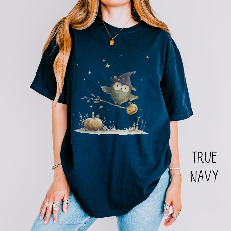 May include: Navy blue t-shirt featuring a whimsical owl wearing a witch hat, perched on a branch with a pumpkin. The design includes stars and a pumpkin. The text "TRUE NAVY" is printed on the right side.