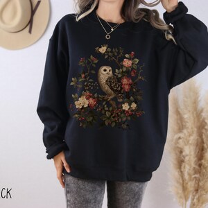Op de afbeelding: Zwart sweatshirt met een gedetailleerde illustratie van een uil omringd door bloemen en bladeren. Het ontwerp bevat bruine, rode en groene tinten. Het woord "BLACK" staat linksonder in de afbeelding.