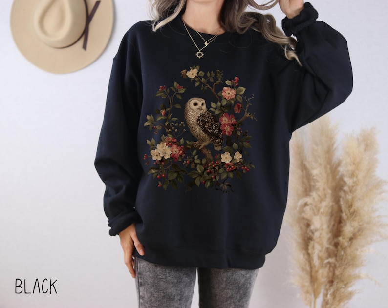 Op de afbeelding: Zwart sweatshirt met een gedetailleerde illustratie van een uil omringd door bloemen en bladeren. Het ontwerp bevat bruine, rode en groene tinten. Het woord "BLACK" staat linksonder in de afbeelding.