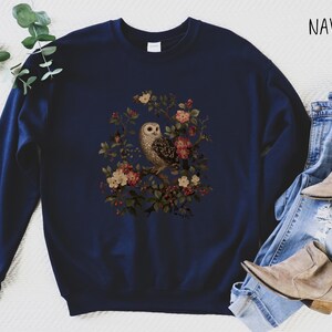 Op de afbeelding: Marineblauwe sweatshirt met een uil en bloemenontwerp. De illustratie toont een uil op een tak met roze, rode en cr&egrave;mekleurige bloemen. Het woord "NAVY" is in het wit rechtsboven gedrukt.