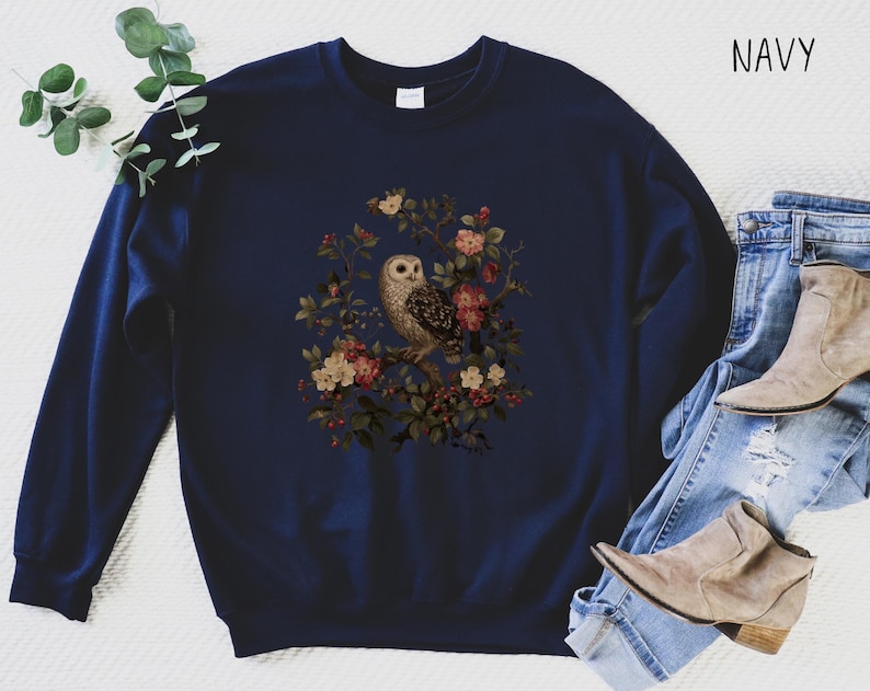 Op de afbeelding: Marineblauwe sweatshirt met een uil en bloemenontwerp. De illustratie toont een uil op een tak met roze, rode en cr&egrave;mekleurige bloemen. Het woord "NAVY" is in het wit rechtsboven gedrukt.