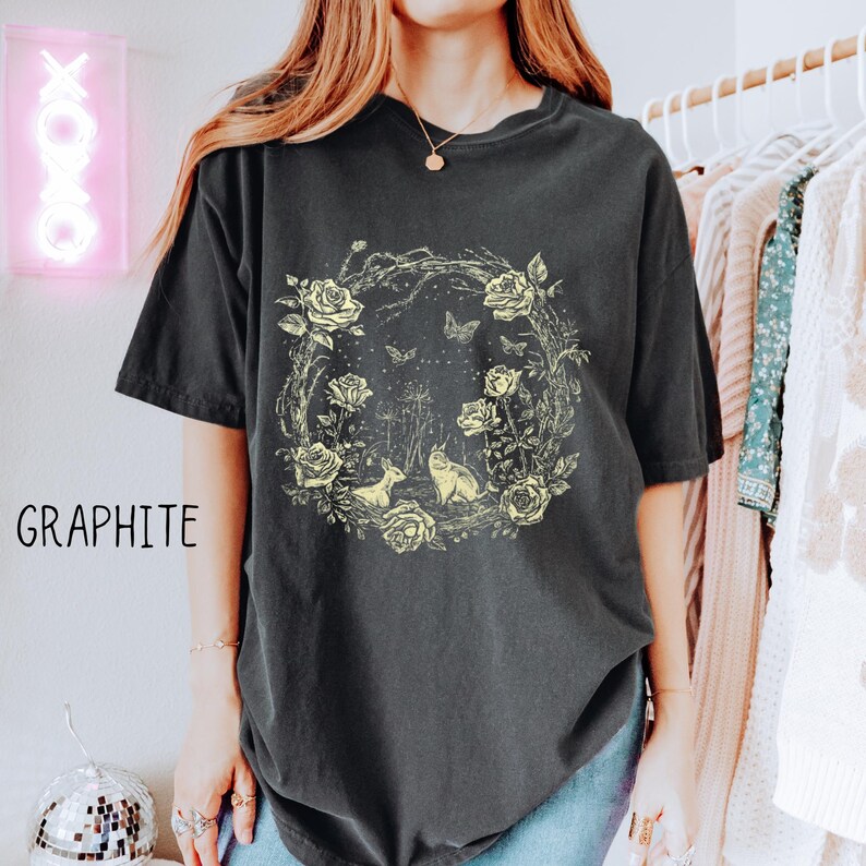 May include: Graphite-colored t-shirt featuring a floral wreath design with roses, butterflies, and two animal figures. The shirt has short sleeves and a relaxed fit. The word "GRAPHITE" is printed on the left side of the shirt.