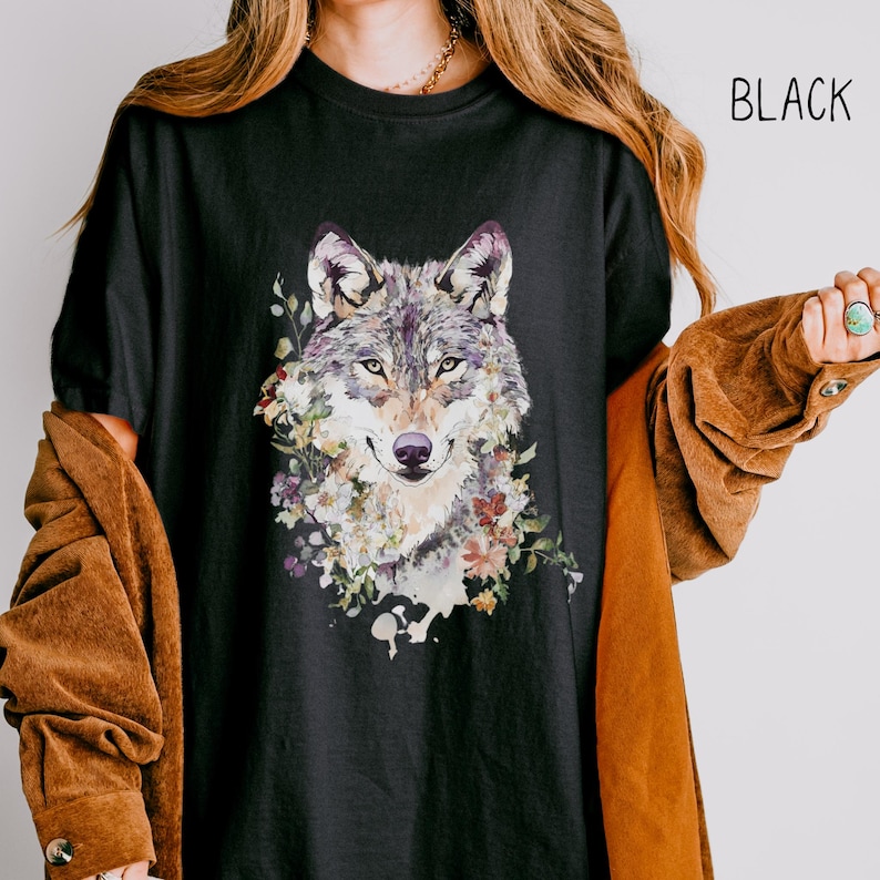 May include: A black t-shirt with a watercolor illustration of a wolf's face surrounded by flowers. The wolf has a gray and brown fur pattern. The flowers are in shades of pink, purple, and yellow. The word 'BLACK' is written in white text in the upper right corner of the image.