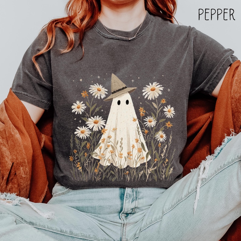 May include: A dark gray t-shirt with a white ghost wearing a brown witch hat in a field of white daisies. The text 'PEPPER' is in the top right corner of the image.