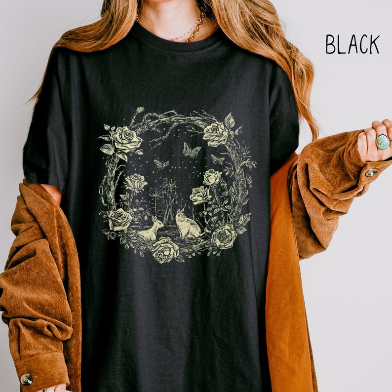 May include: Black t-shirt featuring a floral wreath design with roses, butterflies, and woodland creatures. The text "BLACK" is printed in white on the upper right. The shirt is a comfortable, casual style.