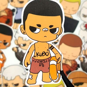 May include: A sticker featuring a cartoon character with tan skin, black hair, and an angry expression. The character wears red swim trunks with the word "KUBO" on the chest and holds an orange life preserver. Other stickers are visible in the background.