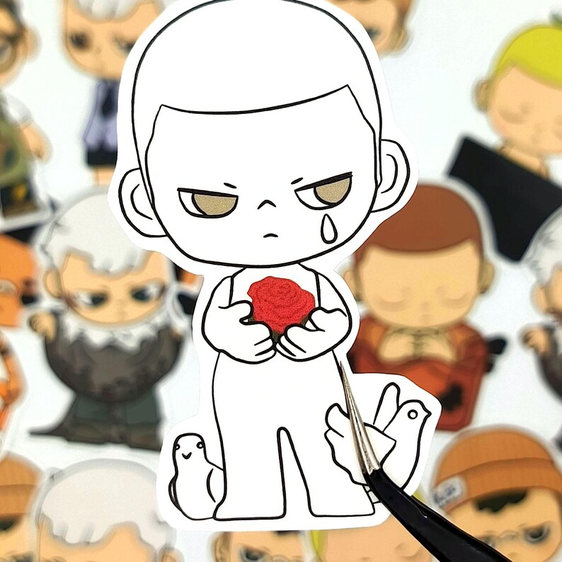 May include: A white sticker featuring a cartoon character holding a red rose. The character has a sad expression with a tear, and is surrounded by other cartoon figures. A pair of tweezers is holding the sticker.