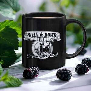 Puede incluir: Taza de cerámica negra con el texto "WELL & DOWD OUTFITTERS" y un gráfico de un ciervo en una mira telescópica. El texto "The buck stops here!" está debajo. Las moras están esparcidas alrededor de la taza.
