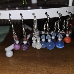 May include: A collection of beaded dangle earrings in various colors, including purple, white, and blue. Each earring features a silver-tone hook and is adorned with multiple beads of different sizes and textures. The earrings are displayed on a white earring stand.