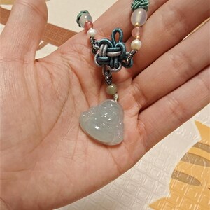 Könnte beinhalten: Eine Halskette mit einem Anhänger aus Jadegrün in Form eines lachenden Buddhas. Die Halskette ist aus einem geknoteten Band gefertigt und hat verschiedene Perlen, darunter rosa, weiße und hellblaue Perlen.