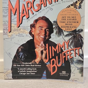 Puede incluir: Portada del libro "Tales from Margaritaville" de Jimmy Buffett. La portada presenta al autor sonriendo, con el título en letras grandes de color naranja. Incluye texto de The New York Times Book Review.