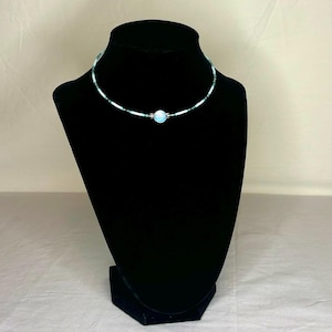 May include: A beaded choker necklace on a black mannequin. The necklace has a central light blue orb, with small white, green, and dark blue beads. The beads are strung together to create a delicate, eye-catching accessory, perfect for adding a touch of elegance.
