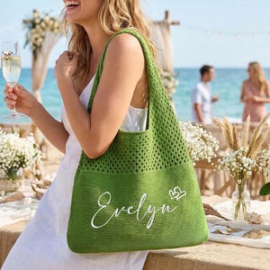 Pu&ograve; includere: Una borsa tote verde all'uncinetto con il nome "Evelyn" in corsivo bianco e due piccoli cuori. La borsa &egrave; tenuta da una persona durante un evento all'aperto vicino all'oceano. La borsa ha un'ampia trama aperta.