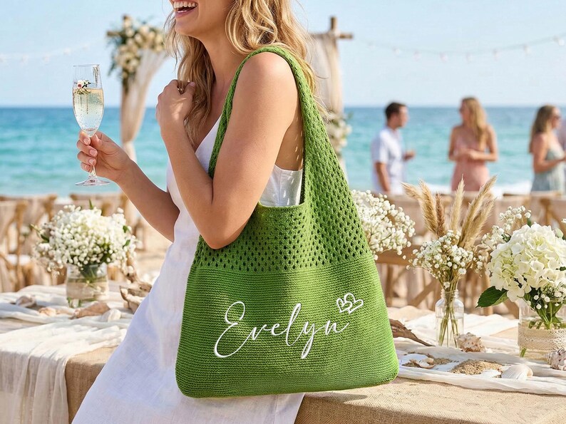 Pu&ograve; includere: Una borsa tote verde all'uncinetto con il nome "Evelyn" in corsivo bianco e due piccoli cuori. La borsa &egrave; tenuta da una persona durante un evento all'aperto vicino all'oceano. La borsa ha un'ampia trama aperta.