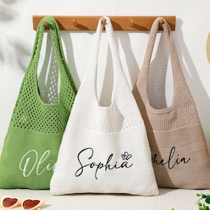 Pu&ograve; includere: Tre borse tote in maglia verde, bianca e beige sono appese a un supporto in legno. La borsa verde ha il nome "Olivia" in corsivo, la borsa bianca "Sophia" e un cuore, e la borsa beige "Amelia". Sono visibili anche un cappello di paglia e occhiali da sole.
