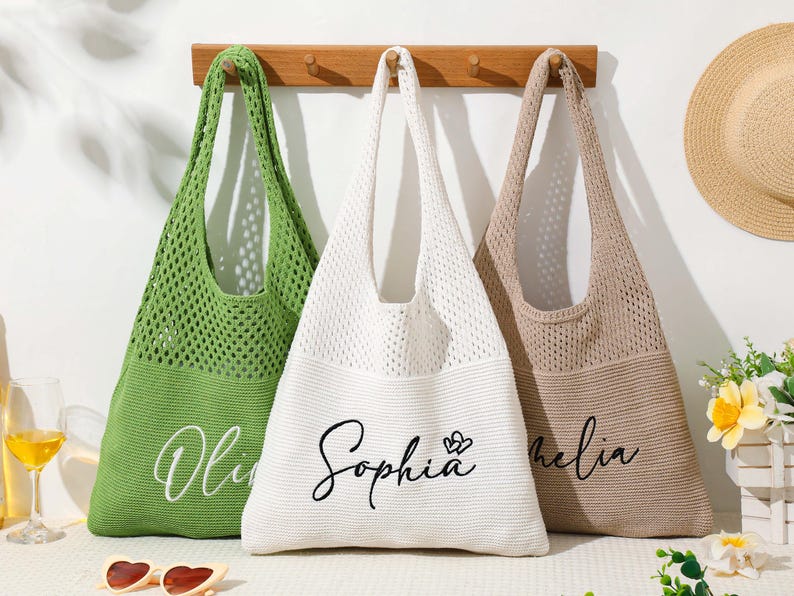 Pu&ograve; includere: Tre borse tote in maglia verde, bianca e beige sono appese a un supporto in legno. La borsa verde ha il nome "Olivia" in corsivo, la borsa bianca "Sophia" e un cuore, e la borsa beige "Amelia". Sono visibili anche un cappello di paglia e occhiali da sole.