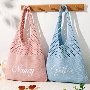 Pu&ograve; includere: Due borse tote personalizzate, una rosa e una blu, sono appese a una parete bianca. Le borse sono realizzate in un materiale a rete lavorato a maglia con fondi pieni. La borsa rosa ha ricamato "Nancy", e la borsa blu ha "Cynthia".