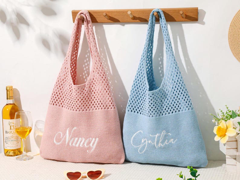 Pu&ograve; includere: Due borse tote personalizzate, una rosa e una blu, sono appese a una parete bianca. Le borse sono realizzate in un materiale a rete lavorato a maglia con fondi pieni. La borsa rosa ha ricamato "Nancy", e la borsa blu ha "Cynthia".