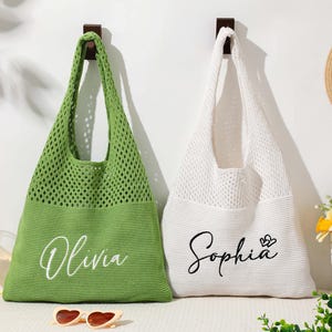 Pu&ograve; includere: Due borse tote in maglia, una verde e una bianca, appese a una parete bianca. La borsa verde ha "Olivia" in corsivo, e la borsa bianca ha "Sophia" con un cuore. Sono visibili anche un cappello di paglia e occhiali da sole.