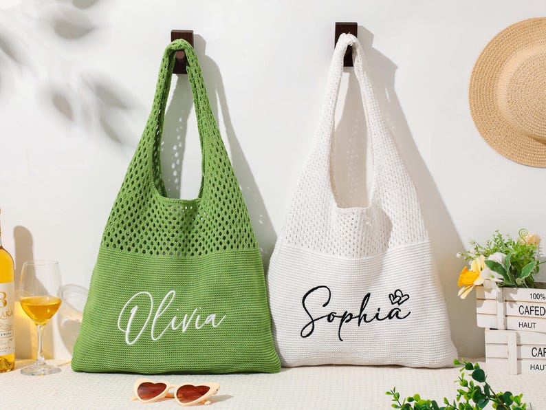 Pu&ograve; includere: Due borse tote in maglia, una verde e una bianca, appese a una parete bianca. La borsa verde ha "Olivia" in corsivo, e la borsa bianca ha "Sophia" con un cuore. Sono visibili anche un cappello di paglia e occhiali da sole.