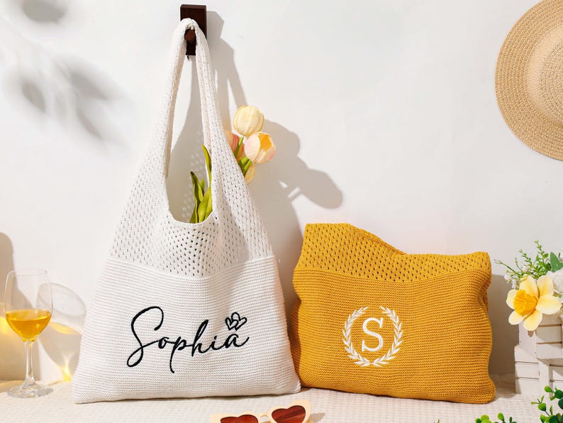Pu&ograve; includere: Una borsa tote bianca a trama aperta con il nome "Sophia" in corsivo, accanto a una pochette gialla con una "S" bianca in una ghirlanda. Un cappello di paglia e fiori sono sullo sfondo.