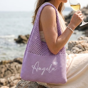 Pu&ograve; includere: Una borsa tote lavanda con un design a rete. La borsa ha un fondo pieno e una tracolla. Il nome "Angela" &egrave; ricamato in bianco sul davanti. Una piccola farfalla bianca &egrave; accanto al nome. Lo sfondo mostra una scena di spiaggia.