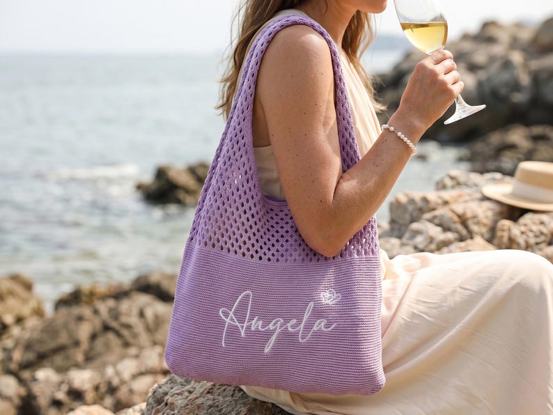 Pu&ograve; includere: Una borsa tote lavanda con un design a rete. La borsa ha un fondo pieno e una tracolla. Il nome "Angela" &egrave; ricamato in bianco sul davanti. Una piccola farfalla bianca &egrave; accanto al nome. Lo sfondo mostra una scena di spiaggia.