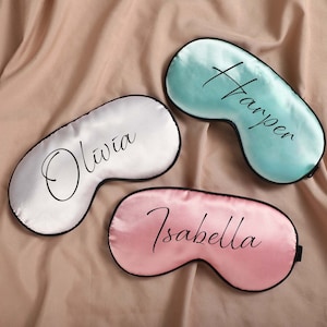 May include: Three satin sleep masks with personalized names. The masks are in white, light blue, and pink.