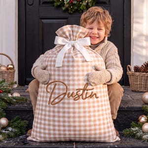 Peut inclure: Un grand sac cadeau en tissu avec un motif vichy beige et blanc. Le sac est resserré en haut par un nœud en ruban blanc et présente le prénom Dustin brodé en lettres cursives marron au centre de la face avant du sac.
