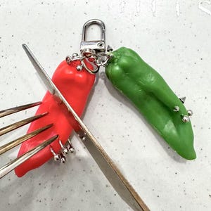 Puede incluir: Un llavero con forma de pimiento rojo y verde conectado por un cierre plateado. El pimiento rojo tiene aros plateados. Un tenedor y un cuchillo se colocan sobre los pimientos.