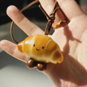 Puede incluir: Un collar con colgante caprichoso con un dije de croissant en miniatura con una cara sonriente y dos pies redondos marrones. El dije está unido a un cordón de cuero marrón, creando un accesorio divertido y único.
