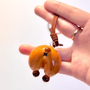 May include: A novelty pendant featuring a glossy, brown, dog butt design with a black cross marking. The pendant is suspended from a brown leather cord. The dog butt has two brown, round, dangling features.