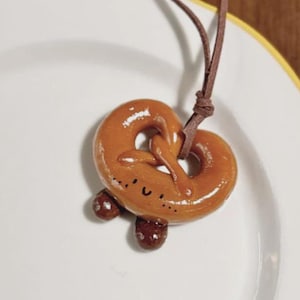 Op de afbeelding: Een bruine pretzel hanger met een leren koord. De hanger heeft een glanzende afwerking en kleine, donkerbruine details. De pretzel ligt op een wit bord met een gele rand.