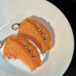Puede incluir: Dos llaveros con forma de salmón naranja con púas plateadas y llaveros. Los llaveros están sobre un plato blanco, con un fondo negro.