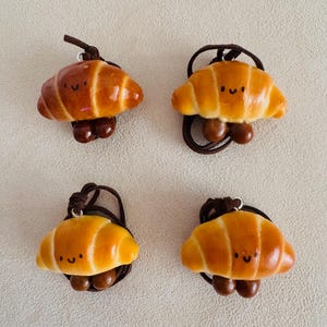 Könnte beinhalten: Vier Miniatur-Croissant-Anhänger mit lächelnden Gesichtern, jeweils an einer braunen Kordel befestigt. Die Croissants sind eine Mischung aus goldbraun und hellgelb, mit dunkelbraunen Akzenten. Die Anhänger sind wahrscheinlich handgefertigt.