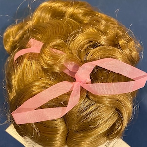 May include: A doll wig with long, wavy, golden blonde hair styled in a bun with two pink ribbons tied around it.