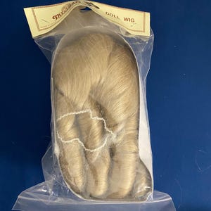 May include: A doll wig in a clear plastic bag. The wig is a light blonde color and has a wavy texture. The packaging has a label that reads "DOLL WIG". The background is a solid blue.