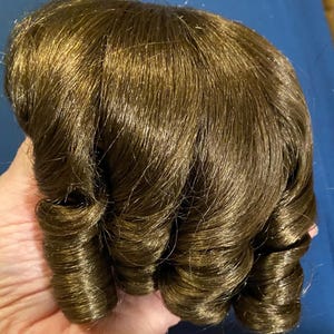 May include: A brown doll wig with long, curled hair. The hair is styled in a series of tight curls.