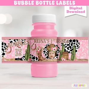 Op de afbeelding: Roze bellenfles met een aangepast etiket. Het etiket heeft een rodeo-thema met een foto van een kind, cowboylaarzen, een hoefijzer en een koeienprint ontwerp. Het etiket leest "Bubbles" en "Reina's 1st Rodeo".