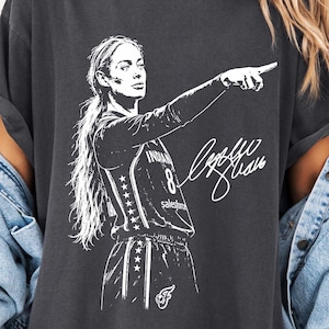 May include: A charcoal gray short-sleeve t-shirt featuring a white graphic illustration of a basketball player pointing forward, with the number 8 and a signature printed on the front. The shirt has a crew neckline and a relaxed, oversized fit.