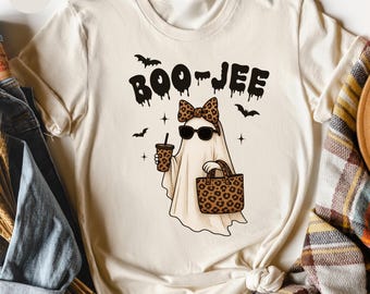 May include: Sand-colored t-shirt featuring a cartoon ghost wearing sunglasses and a leopard print bow, holding a drink and a matching tote bag. The text "BOO-JEE" is above the ghost, with bats and stars. A casual, fun Halloween design.