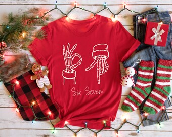 May include: A red t-shirt with a white graphic of skeleton hands forming the numbers six and seven. The text "Six Seven" is written below the hands. The shirt is part of a Christmas-themed display with a stocking, jeans, and festive decorations.