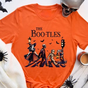 May include: Orange t-shirt with a Halloween-themed graphic. The design features a witch, skeleton, and mummy crossing a street, with the text "The Boo-tles" above. Decorative spiders and bats are also present.